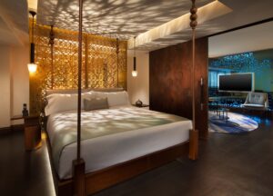 Four-poster bed in a modern bedroom with patterned ceiling shadows, adjacent to a living area with a TV.