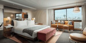 A modern bedroom with a large bed, pink foot bench, and orange leather window seating overlooking a cityscape through slatted blinds.
