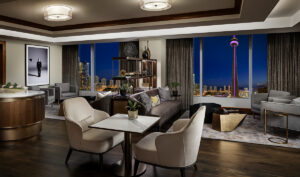 Modern suite living room with dark wood flooring, multiple seating areas, and floor-to-ceiling windows displaying the CN Tower at night.