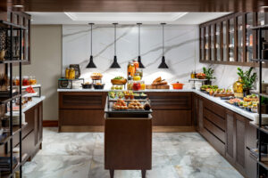 A buffet station with white marble floors and walls, dark wood cabinets, and four black pendant lights over prepared food displays.