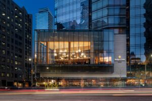 Exterior view of a modern glass hotel at dusk, with a brightly lit interior floor showing multiple chandeliers and car light trails.