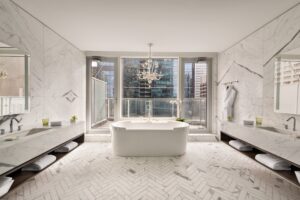 Luxury bathroom with white freestanding tub before city view windows, flanked by marble double vanities and herringbone floor.