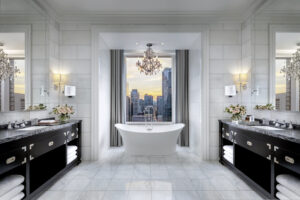 Symmetrical bathroom with light marble walls and flooring. A white soaking tub faces a cityscape view, flanked by two black double vanities.