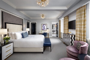 A well-appointed bedroom with a large bed, blue desk chair, television, patterned curtains, and visible bathroom.