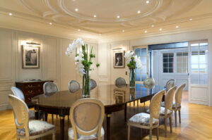 A luxurious dining room with a long polished table, ten upholstered chairs, paneled walls, and an ornate ceiling.