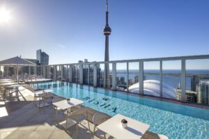 Rooftop pool with city skyline, CN Tower, and lake under bright sun. White tables, chairs, and loungers surround the pool.