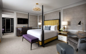 A luxurious bedroom with a yellow four-poster bed, wall-mounted TV, patterned armchair, and crystal chandelier.