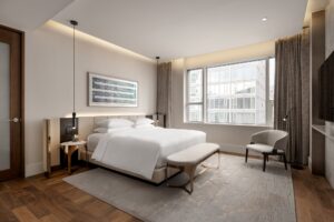 A modern bedroom with a king-sized bed, a large window overlooking city buildings, and neutral-toned furniture.