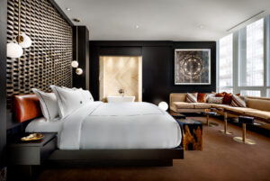 Modern bedroom with dark bed, white bedding, freestanding tub behind sliding doors, L-shaped sofa, and large windows.