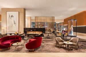 Hotel lobby lounge with red velvet and gray seating, black marble fireplace, patterned rug, and display cases.