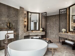 Modern bathroom with a large freestanding tub, double vanity sinks, mirrors, stone walls, and bronze-toned fixtures.