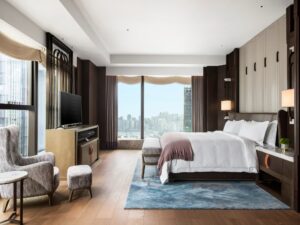 Modern bedroom with large bed, city skyline view, armchair, TV, wooden floors, and blue geometric rug.