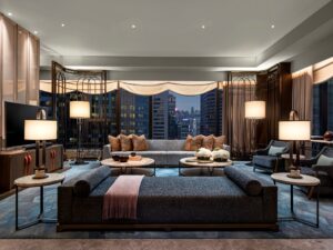 A luxury living room with sofas, armchairs, and coffee tables, featuring city views from large windows at night.
